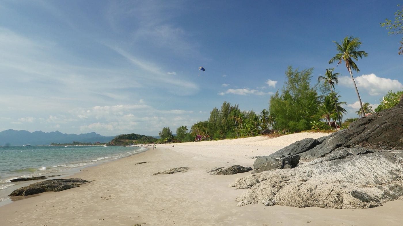 Wide sandy beach on Langkawi with palm trees, dark rocks and a parasail overhead
